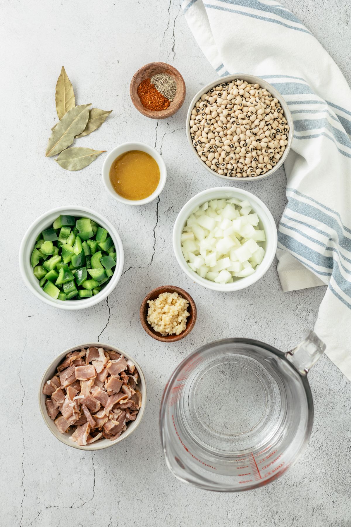 Ingredients for Black-eyed Peas Dish in separate bowls: Diced bacon, yellow onions, green bell pepper, minced garlic, dried black-eyed peas, water, chicken bouillon, bay leaves, black pepper, Cajun seasoning