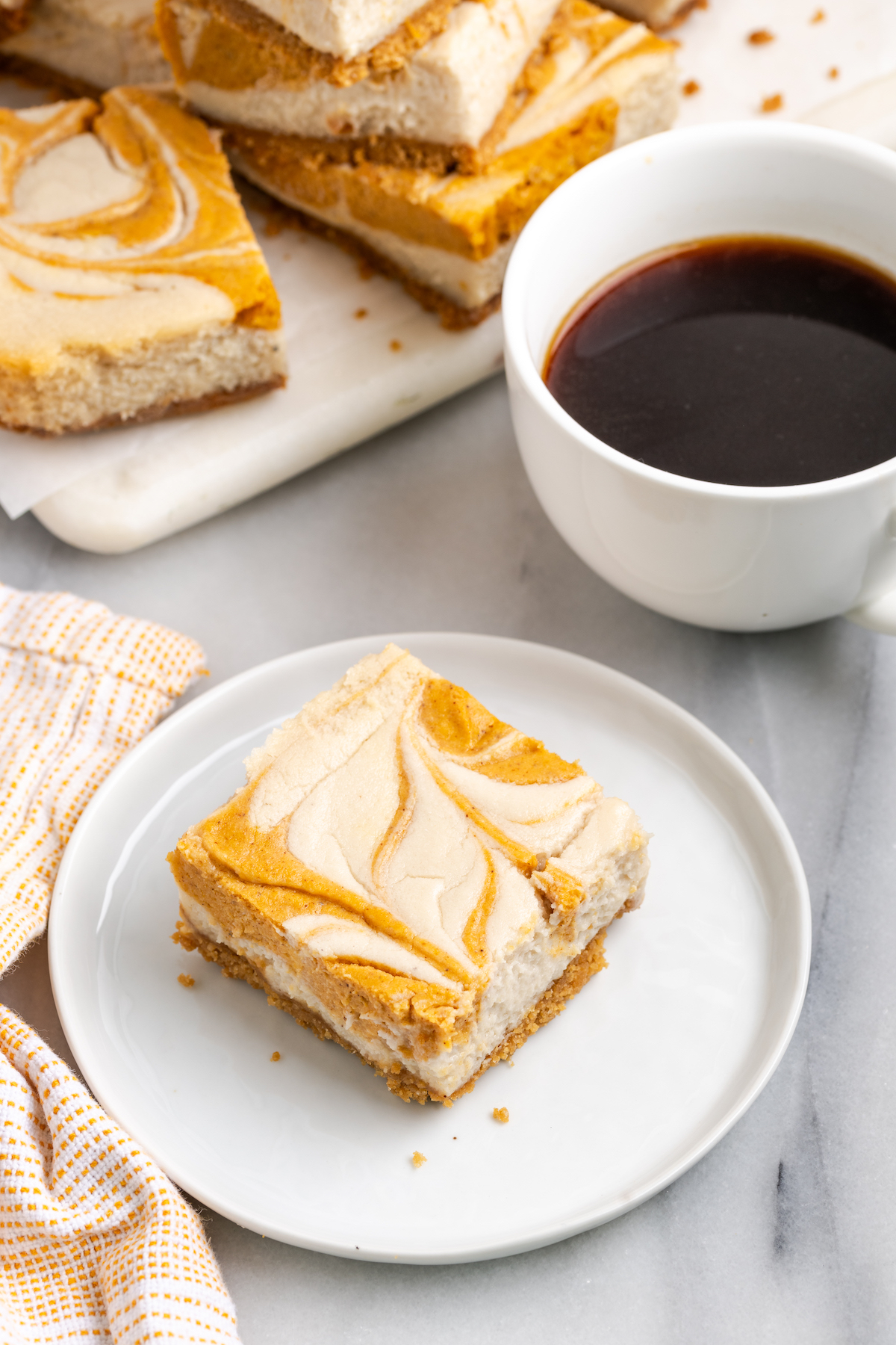 pumpkin pie cheesecake bar on plate next to cup of coffee and additional bars on cutting board
