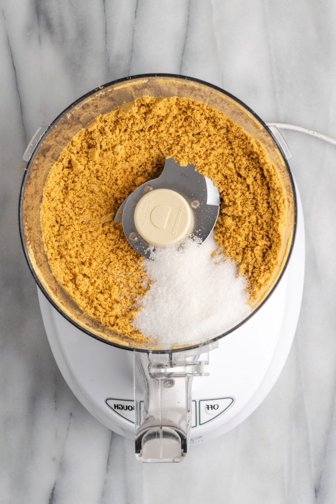 Overhead view of sugar and salt added to graham crumbs in food processor