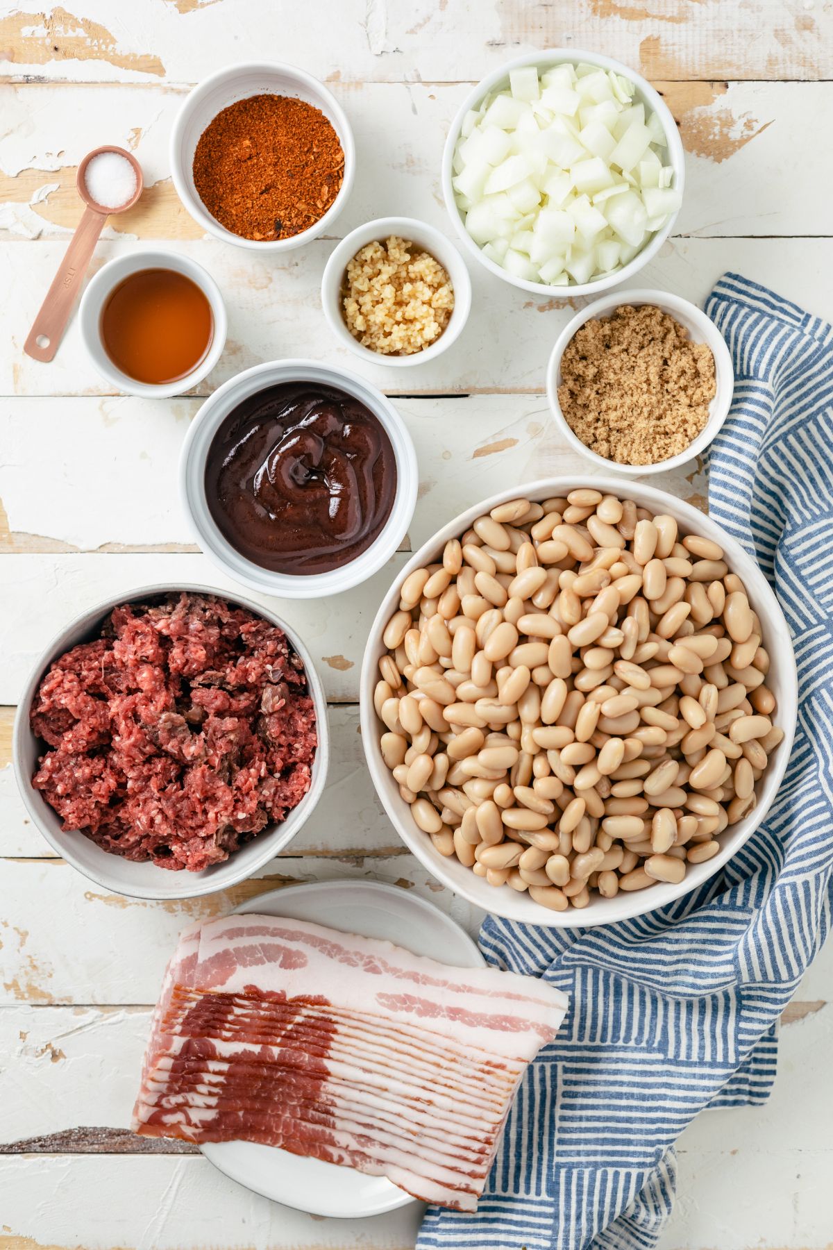Ingredients for savory baked beans: no sugar-added bacon, diced yellow onion, minced cloves, lean ground beef or turkey, cannellini beans, BBQ seasoning, hickory or Kansas City style BBQ sauce, brown sugar, Hennessy (or your preferred cola or broth), and sea salt, each in its own bowl.