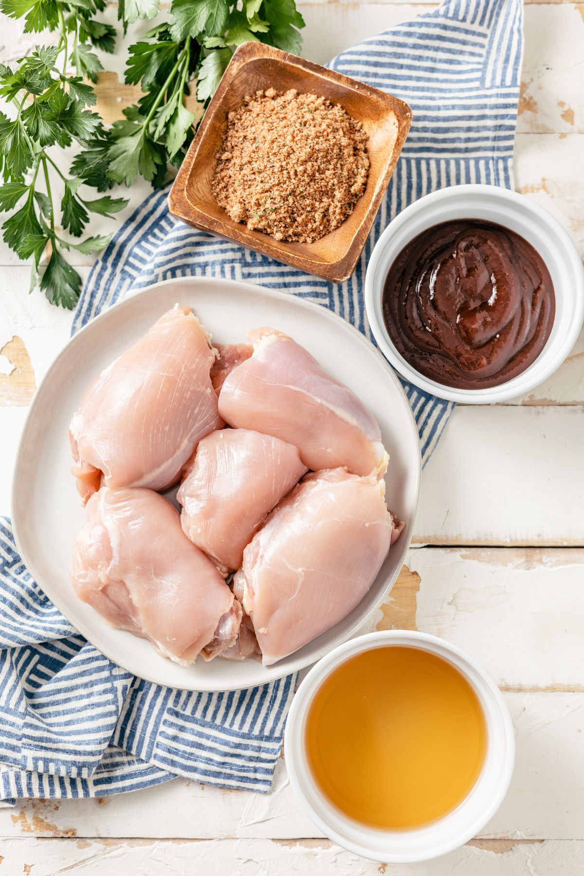 ingredients for pulled bbq chicken in separate bowls containing boneless, skinless chicken thighs, BBQ chicken rub, chicken broth, and hickory BBQ sauce.