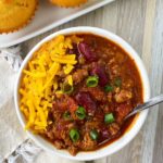 Beef Chili in a white bowl with a spoon, garnished with green onions, cheese next to a plate of cornbread muffins