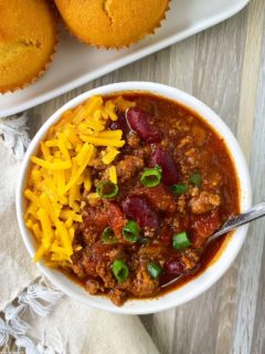 Beef Chili in a white bowl with a spoon, garnished with green onions, cheese next to a plate of cornbread muffins