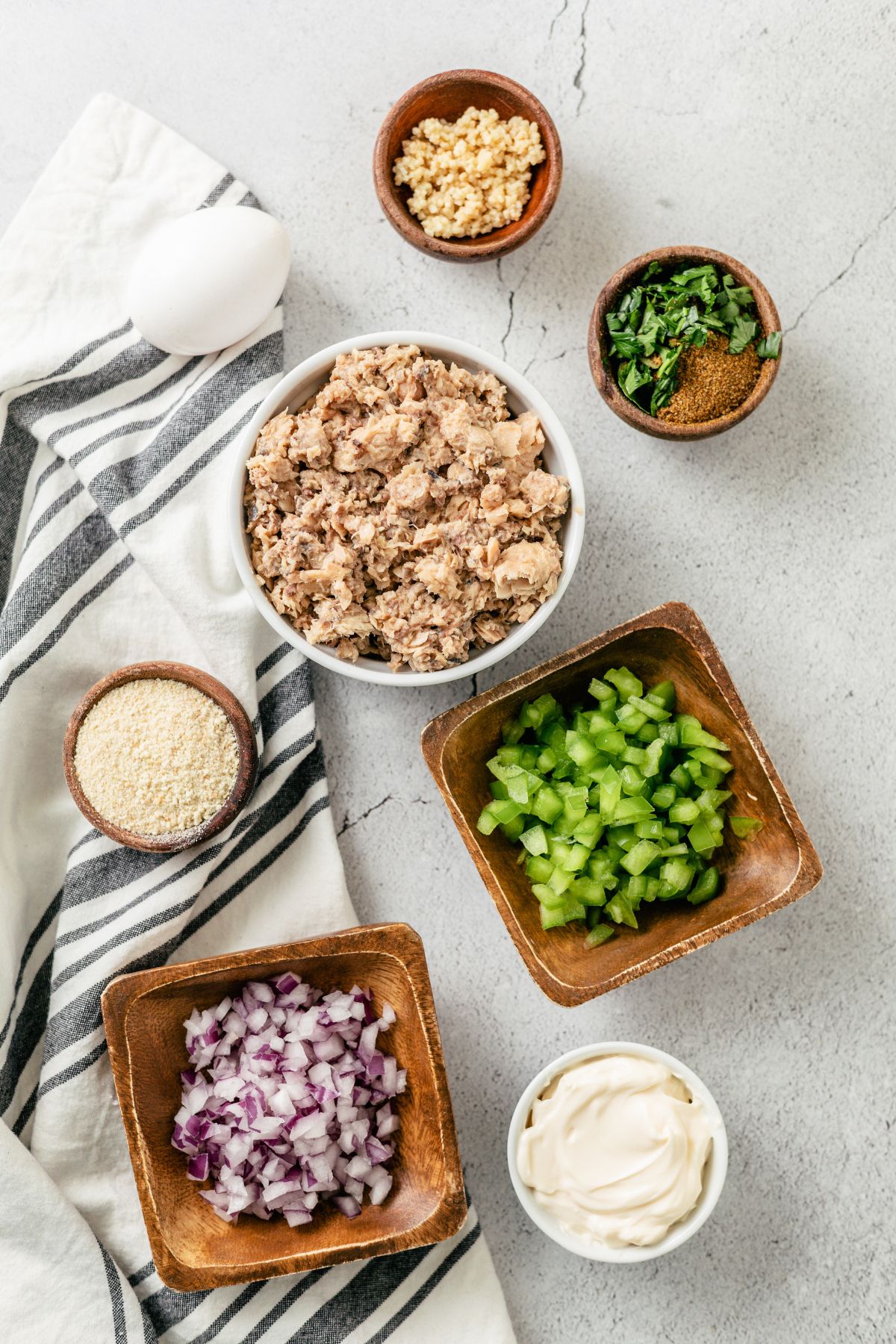 Ingredients for salmon patties with canned salmon in separate bowls: canned salmon, shallots, green bell peppers, garlic, mayo, egg, parsley, Old Bay seasoning, and breadcrumbs.