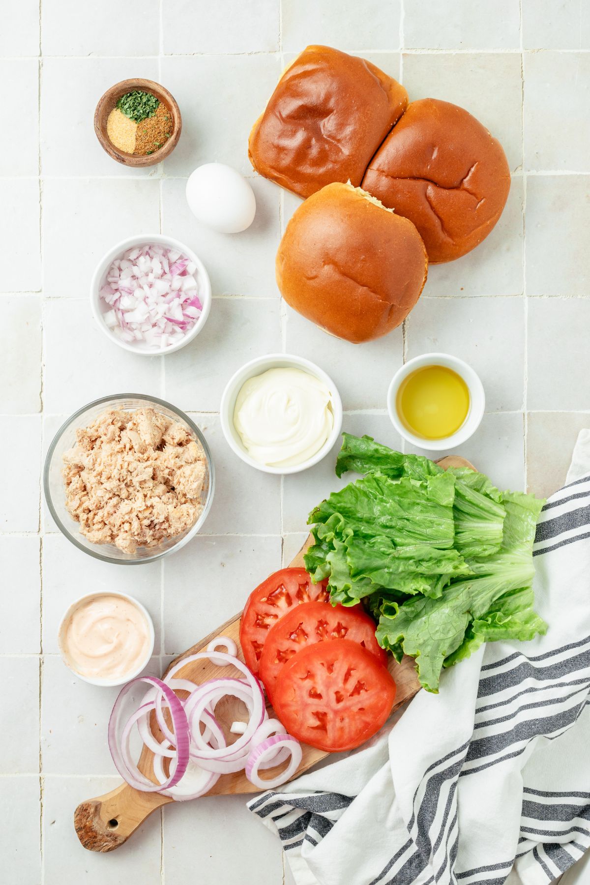 canned salmon burger ingredients arranged in separate bowls: boneless skinless canned salmon, finely chopped red onions, garlic powder, full-fat mayo for moistness, an egg, dried parsley, Old Bay seasoning, avocado oil for sautéing, and brioche burger buns