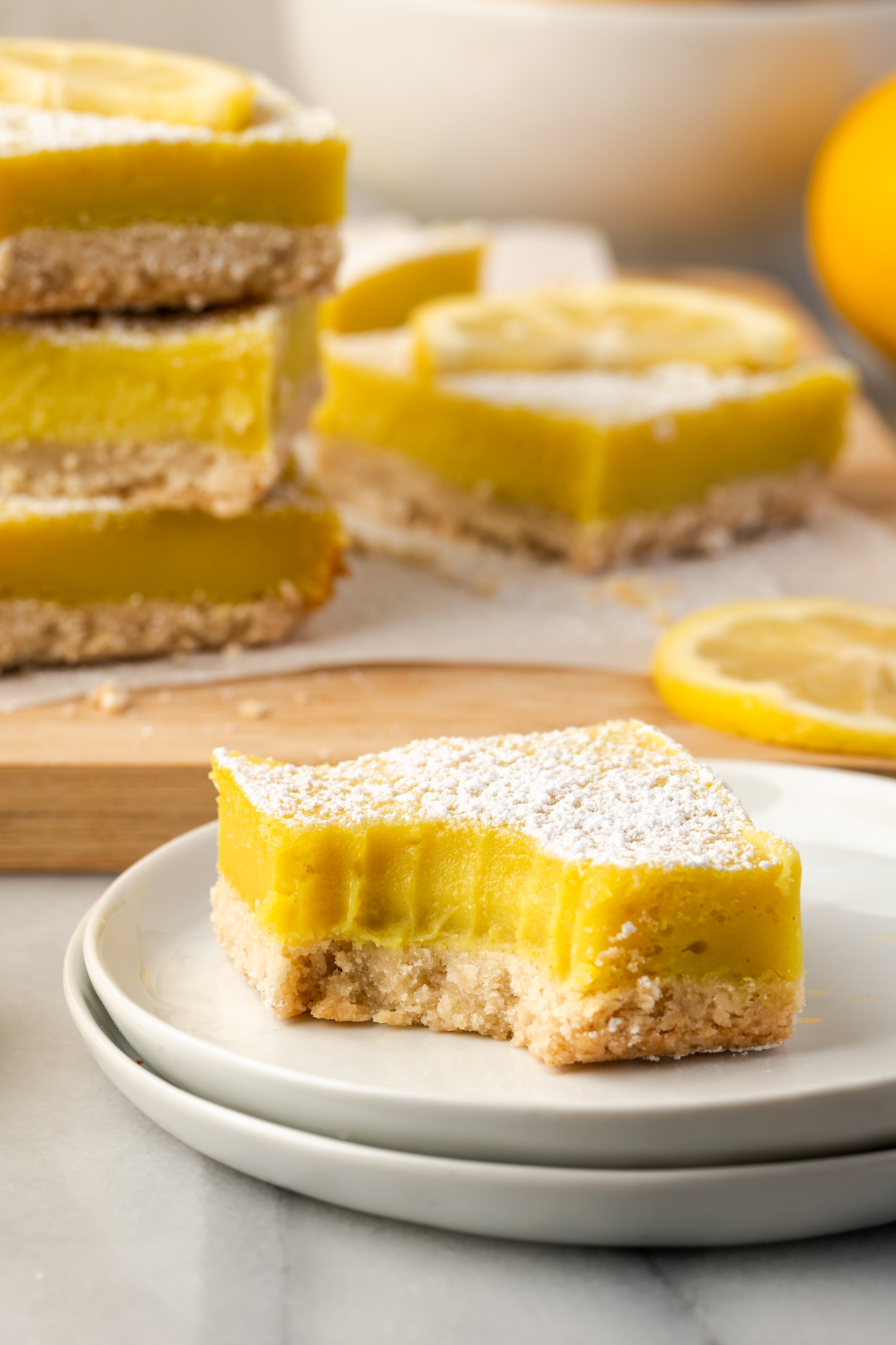 Bitten lemon bar on plate with stack of lemon bars on board in background